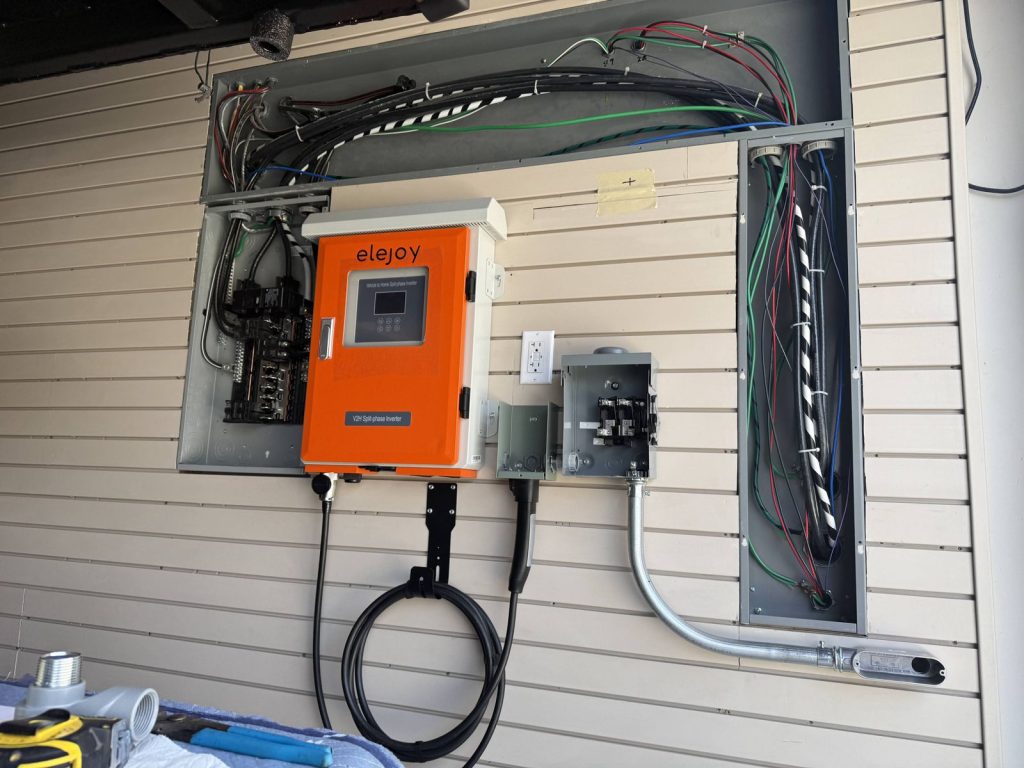 Elejoy V2H inverter mounted on a garage wall beside the subpanel, with its charging cable on a holster, a manual transfer switch and conduit to the main panel
