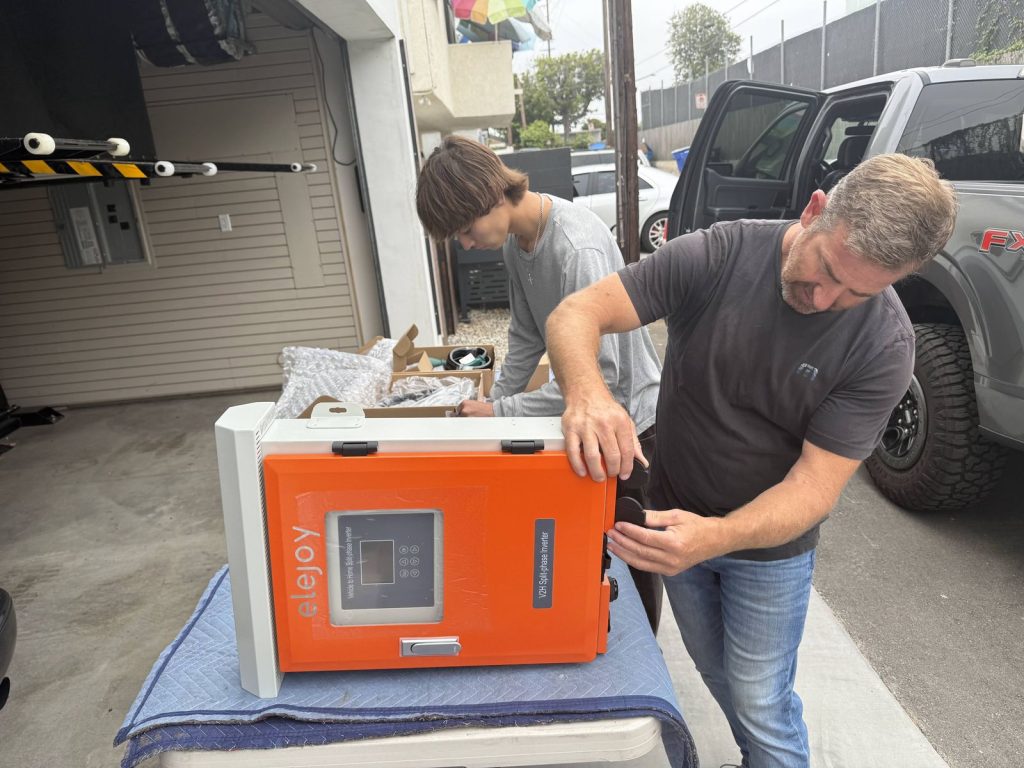 Two people unboxing the orange Elejoy V2H split-phase inverter on a workbench in a driveway